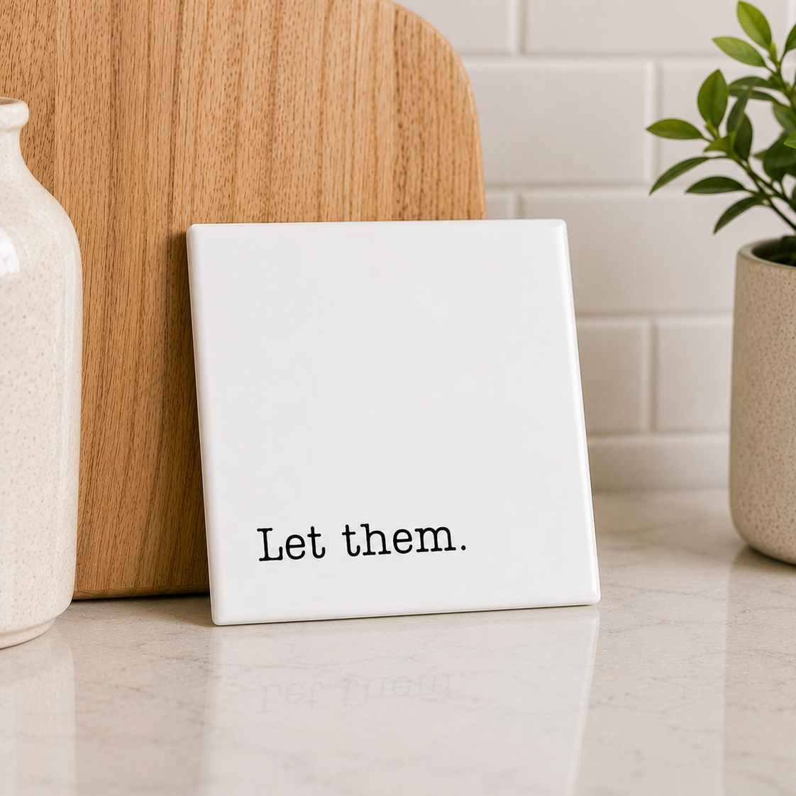 White square tile with 'Let them.' text on a kitchen counter with decor items.