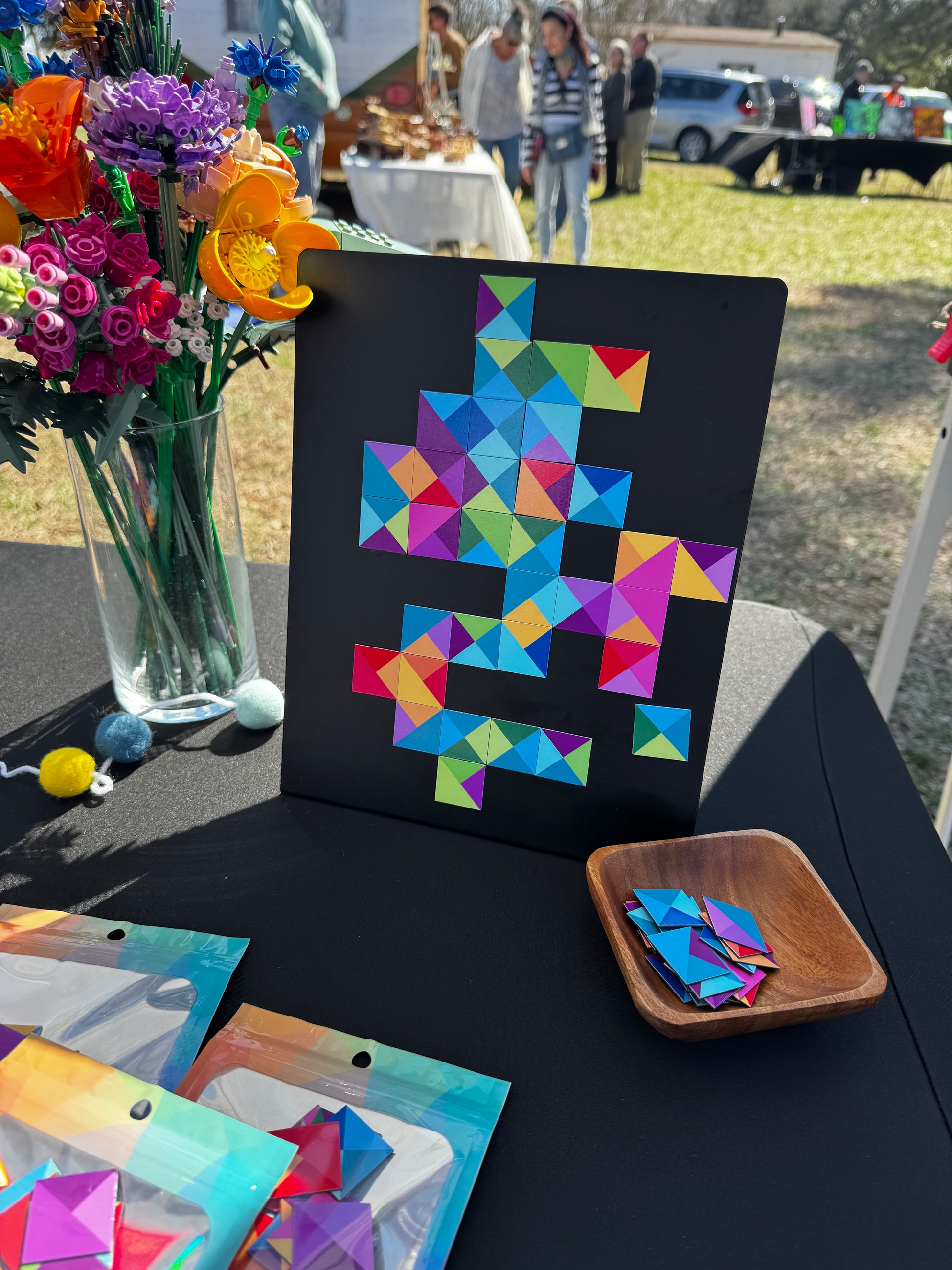 Another angle of a display of magnetic mosaic tiles at a vendor market with a magnet board and pieces for customers to test, and packaged tiles on the table in colorful pouches