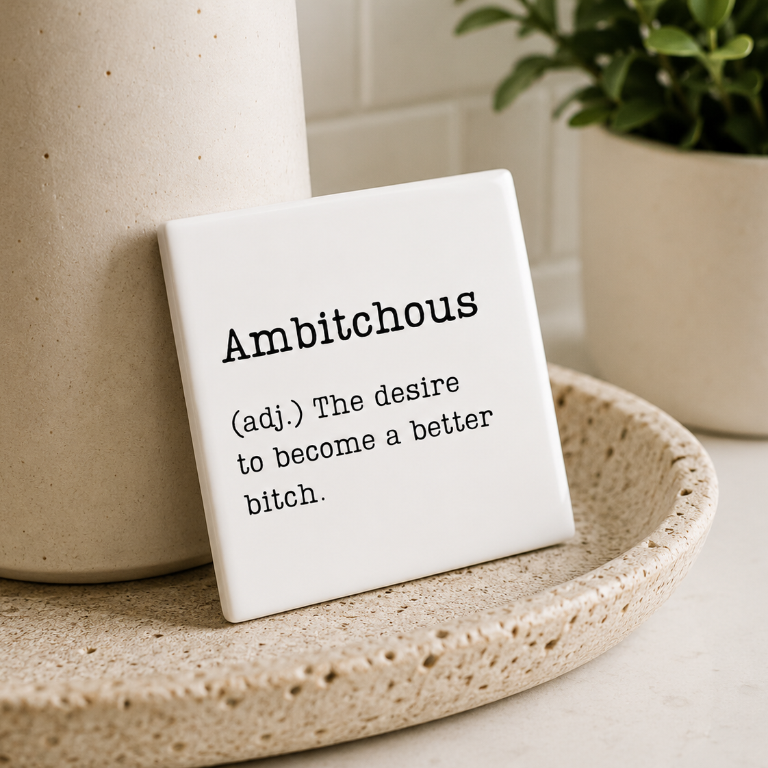 Small decorative tile with the word 'Ambitchous' on a ceramic plate with a plant in the background.